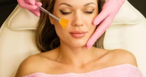 Woman receiving a chemical peels treatment, eyes closed and relaxed, while a professional in pink gloves applies the solution to her cheek with a fan brush in Evans, GA